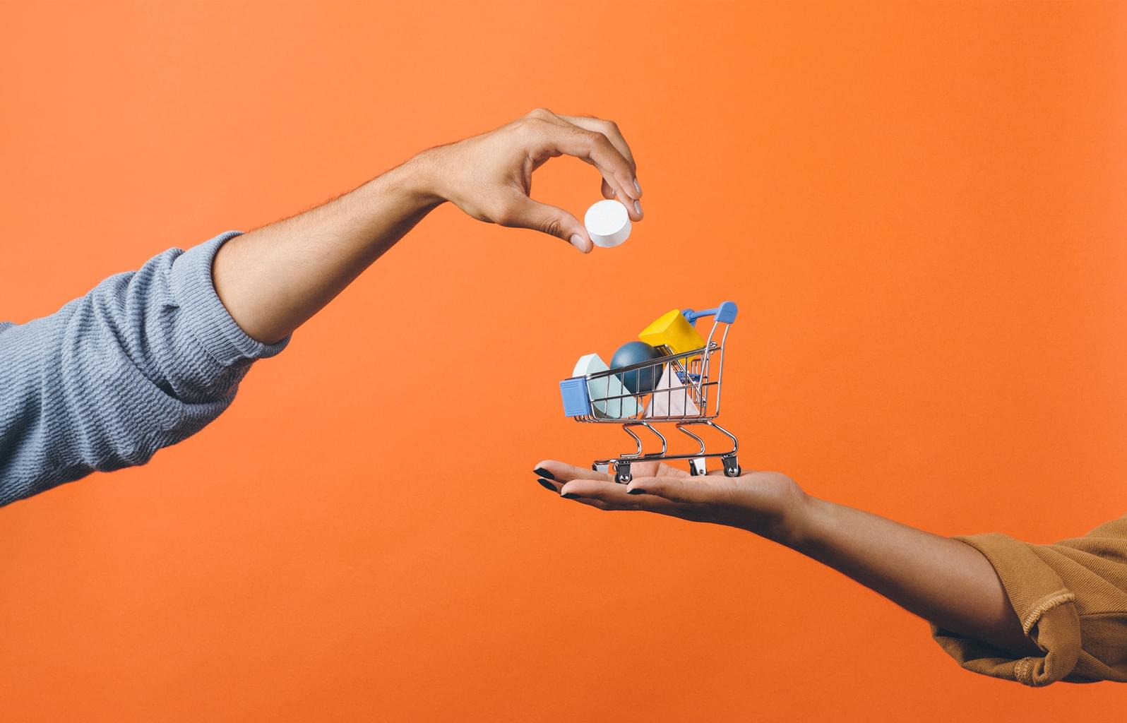 Person holding a tiny shopping cart in the palm of their hand. The shopping cart is full of small objects, and someone is adding another small object to the cart